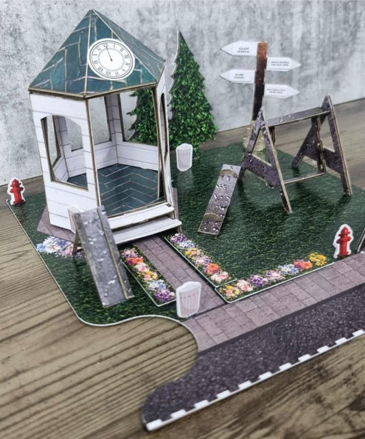 Detailed paper diorama of a community park with gazebo, swing set, flower-lined path, fire hydrants, and directional signs.