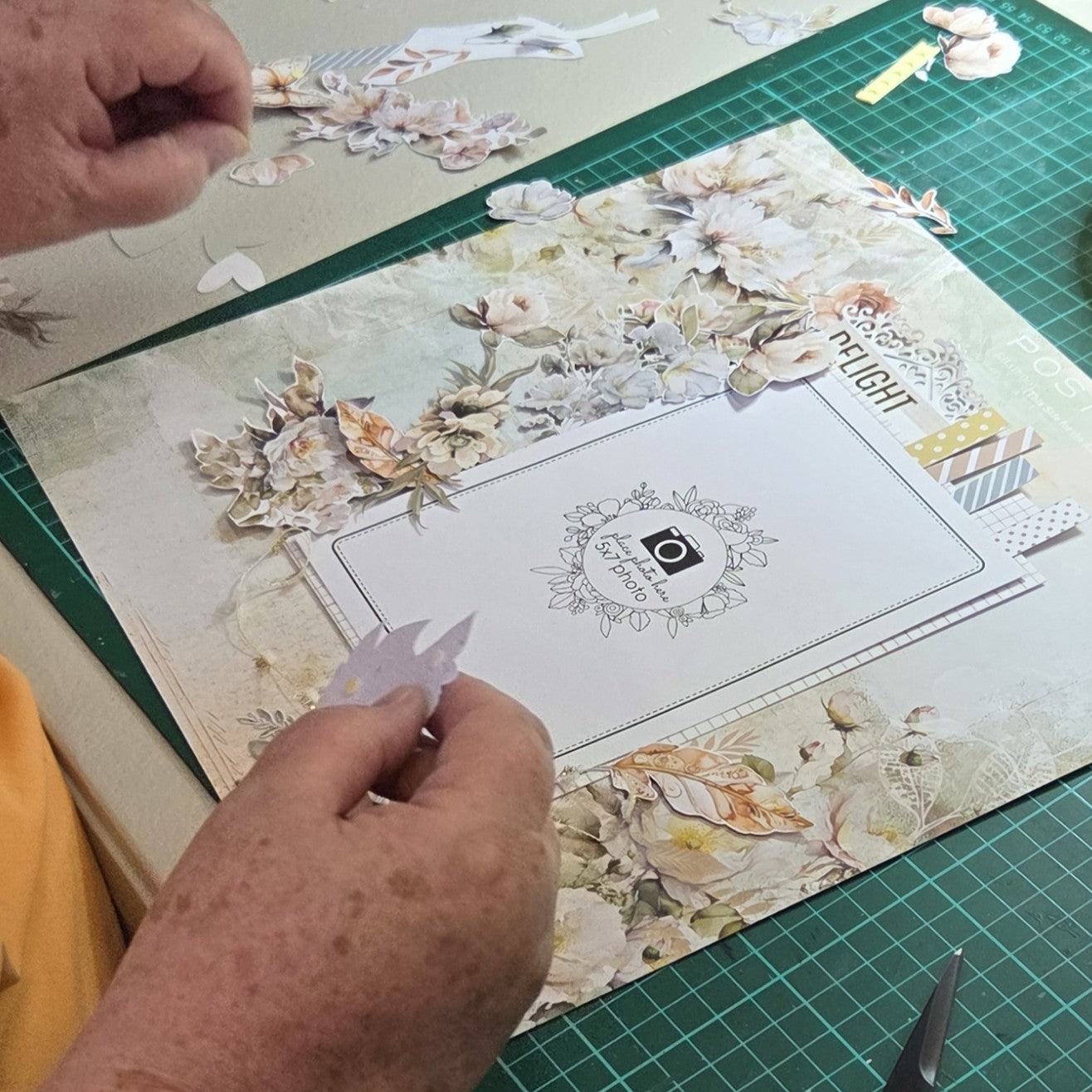 Close-up of hands arranging die-cut floral elements on a scrapbook layout with vintage botanical papers and journaling space