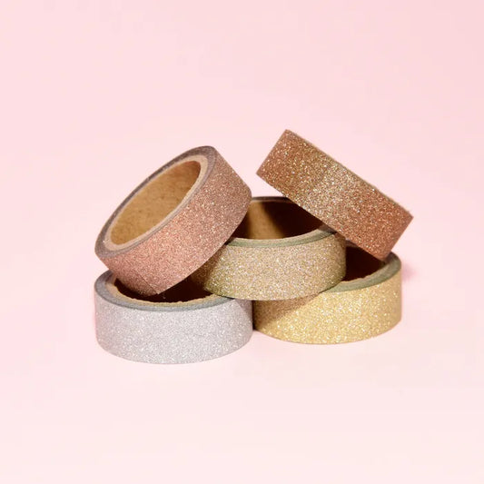 Five rolls of glitter tape in gold, rose gold, and silver on a pink background.