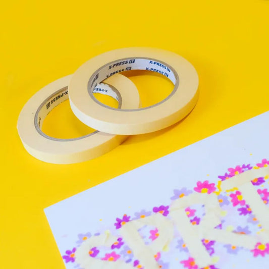 Two rolls of masking tape on a yellow background with a piece of paper featuring colourful designs showing how they have masked off an area to decorate around it.