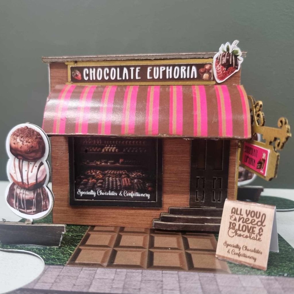 Front view of the Chocolate Euphoria shop with display window, All You Need is Love and Chocolate sandwich board sign, and jigsaw base board