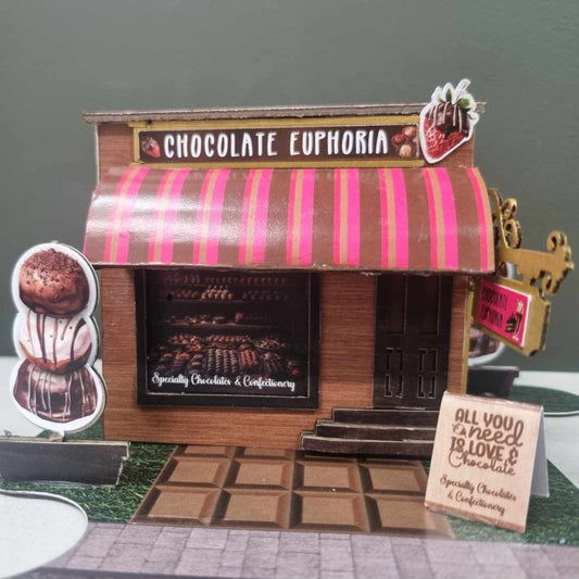 Front view of the Chocolate Euphoria shop with display window, All You Need is Love and Chocolate sandwich board sign, and jigsaw base board
