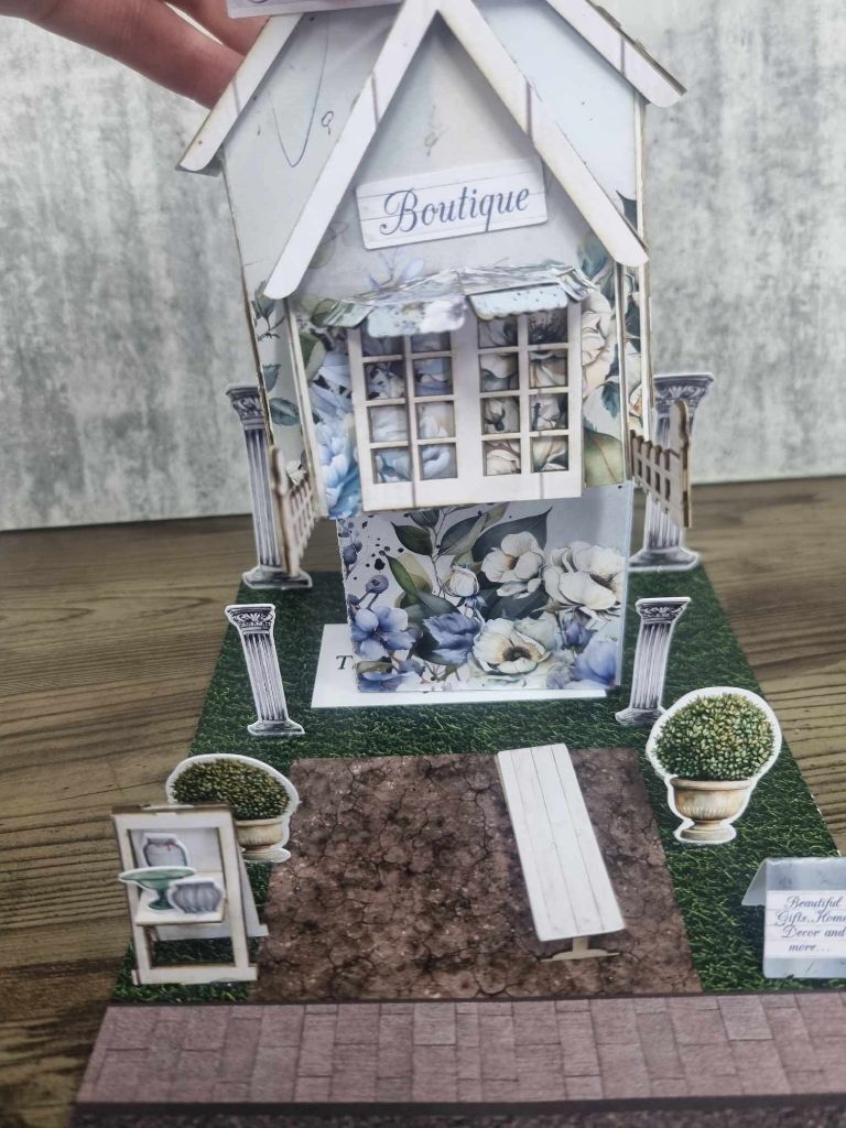 3Quarter Designs Enchanting Treasures Boutique Gift Shop miniature building being lifted to reveal a hidden mini album underneath, decorated with blue floral botanical papers and topiary urns on the base board