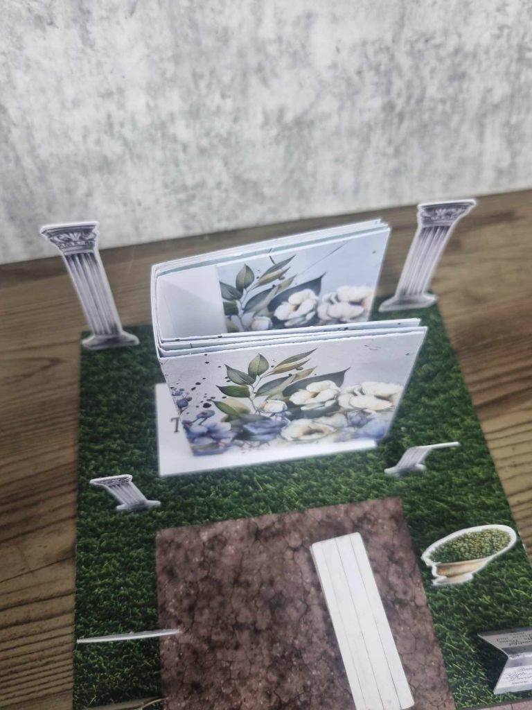 3Quarter Designs Enchanting Treasures Boutique Gift Shop hidden mini album sitting open between the ornate columns on the grass base board, decorated with blue and white floral botanical papers