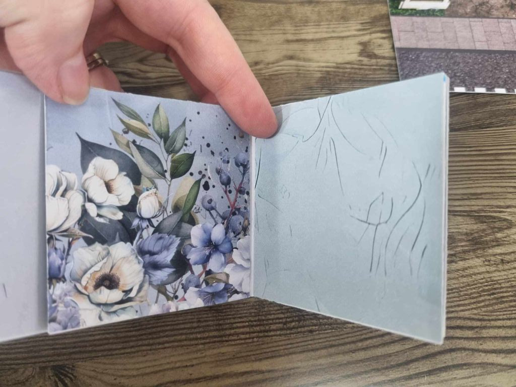 3Quarter Designs Enchanting Treasures Boutique Gift Shop hidden mini album open showing an interior page decorated with watercolour blue and white florals including roses, hydrangeas, and botanical foliage, alongside a plain soft blue journaling page