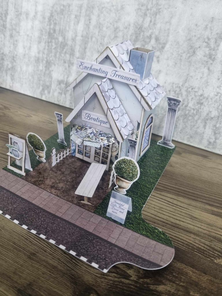 3Quarter Designs Enchanting Treasures Boutique Gift Shop miniature building angled front view showing scalloped roof tiles, chimney, arched side window, ornate columns, topiary urns, and jigsaw base board