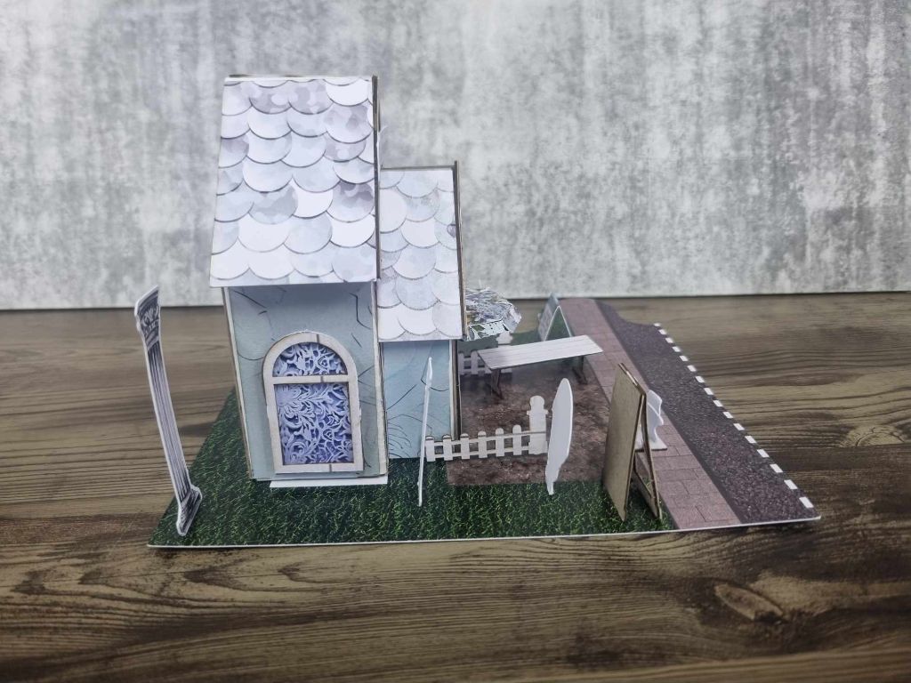3Quarter Designs Enchanting Treasures Boutique Gift Shop miniature building left side view showing scalloped roof tiles, arched window with blue lace detail, picket fence, pergola, ornate column, and jigsaw base board with grass and cobblestone road section