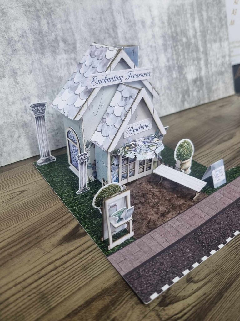 3Quarter Designs Enchanting Treasures Boutique Gift Shop miniature building angled front left view showing Enchanting Treasures sign, Boutique entrance, scalloped roof, ornate column, arched window with blue lace detail, topiary urn, display frame, and A-frame sign on cobblestone path
