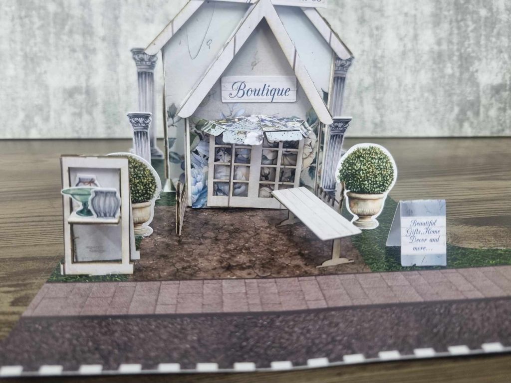 Close-up of 3Quarter Designs Enchanting Treasures Boutique Gift Shop miniature building showing Boutique sign, ornate columns, display window with gift items, topiary urns, display frame with vase artwork, bench seat, and A-frame sign reading Beautiful Gifts Home Decor and more