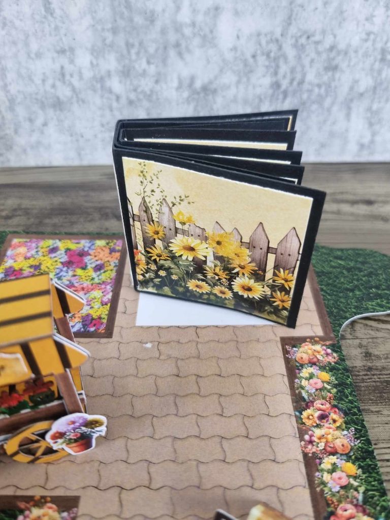 Mini album standing open on the cobblestone path in front of the florist, showing the sunflower picket fence cover