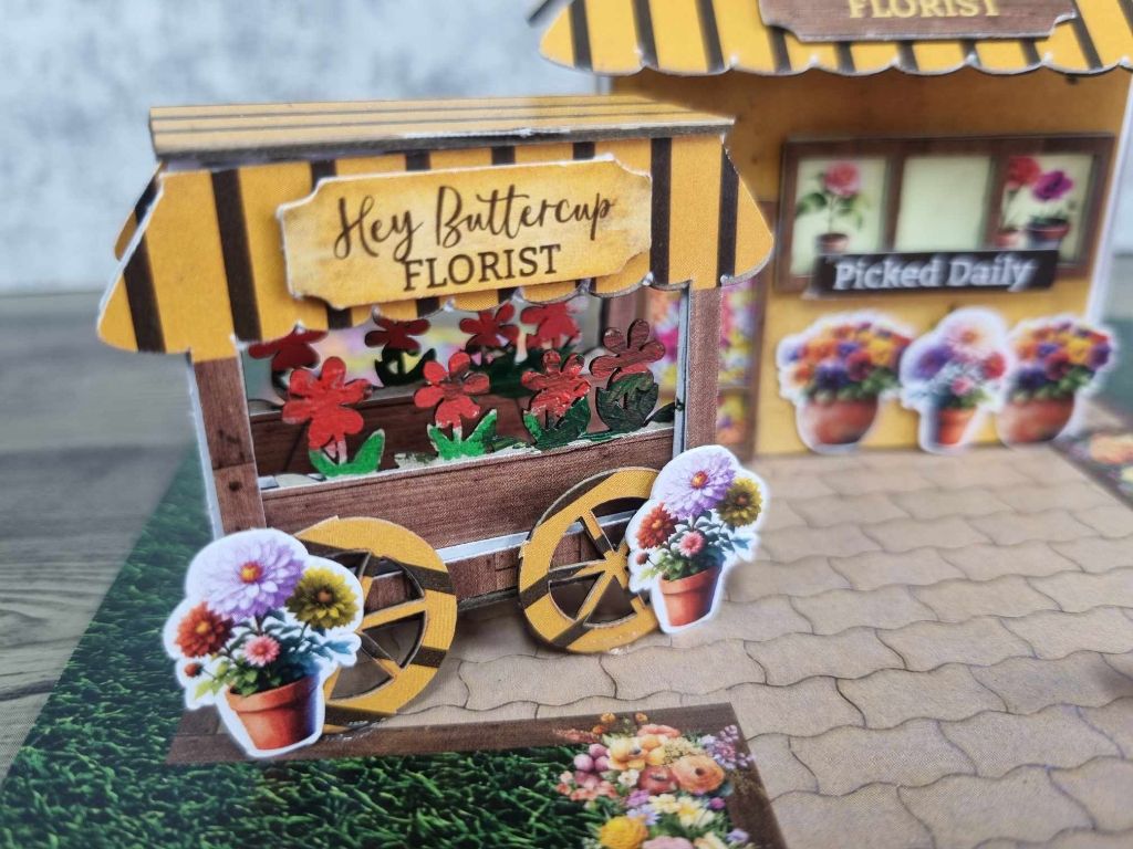 Assembled Hey Buttercup Florist miniature building with sunny yellow facade, black and gold striped awning, and flower cart with wooden wheels