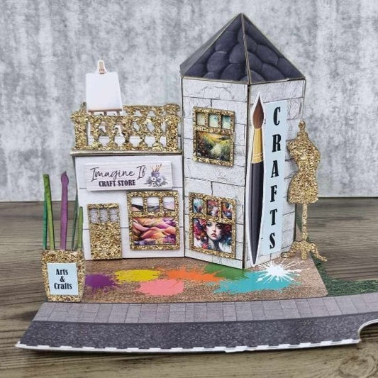 3Quarter Designs Imagine If Craft Store miniature building front view showing white crackle wood panel walls, Imagine If Craft Store sign, gold glitter ornate window frames with art print displays, paintbrush and CRAFTS tower panel, gold glitter dressmaker's dummy die-cut, easel, coloured pencil die-cuts, Arts and Crafts sign, and colourful paint splash base board with cobblestone road section