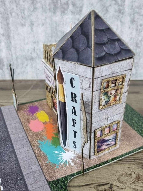 3Quarter Designs Imagine If Craft Store miniature building angled front-right view showing paintbrush and CRAFTS tower panel, dark hexagonal slate roof, gold glitter ornate window frames with art print displays, colourful paint splash base board, and cobblestone road section