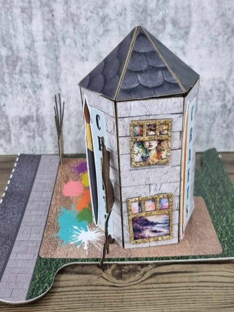 3Quarter Designs Imagine If Craft Store miniature building rear-right view showing the full hexagonal tower with dark slate pyramid roof, crackle white wood panel walls, gold glitter framed art print windows on two faces, paint splash base board, and grass jigsaw base board with cobblestone road section