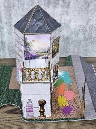 3Quarter Designs Imagine If Craft Store miniature building left side view showing hexagonal tower with dark slate pyramid roof, large landscape art print window with gold glitter frame, gold glitter ornate balustrade, ink bottle and rubber stamp die-cuts at ground level, and colourful paint splash base board with grass jigsaw section