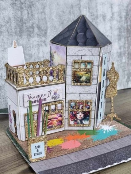 3Quarter Designs Imagine If Craft Store miniature building angled front-left view showing Imagine If Craft Store sign, gold glitter ornate window frames with art print displays, easel with blank canvas, gold glitter balustrade, Arts and Crafts sign, CRAFTS lettering with gold glitter dressmaker's dummy die-cut, hexagonal tower with dark slate pyramid roof, and colourful paint splash base board