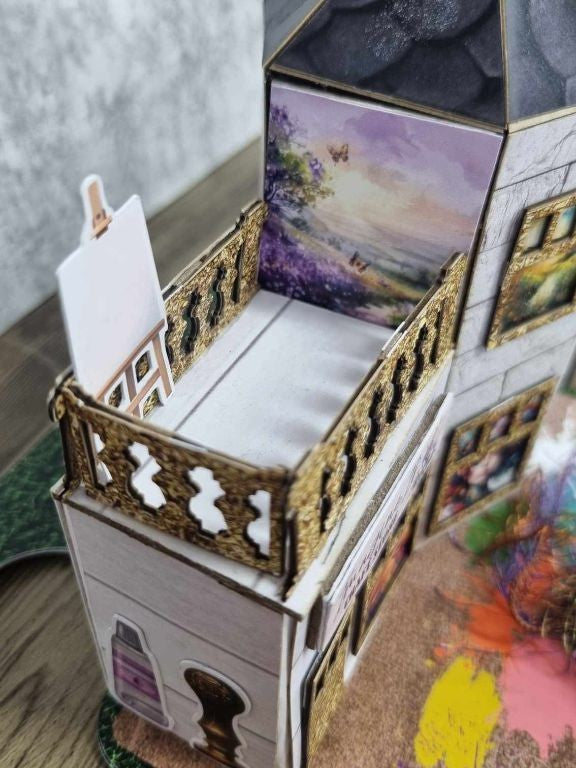 Close-up of 3Quarter Designs Imagine If Craft Store miniature building balcony showing intricate gold glitter ornate balustrade, mini album pages tucked inside the balcony area, easel with blank canvas die-cut, purple landscape art print wrapping around the upper tower panels, and gold glitter framed art print windows