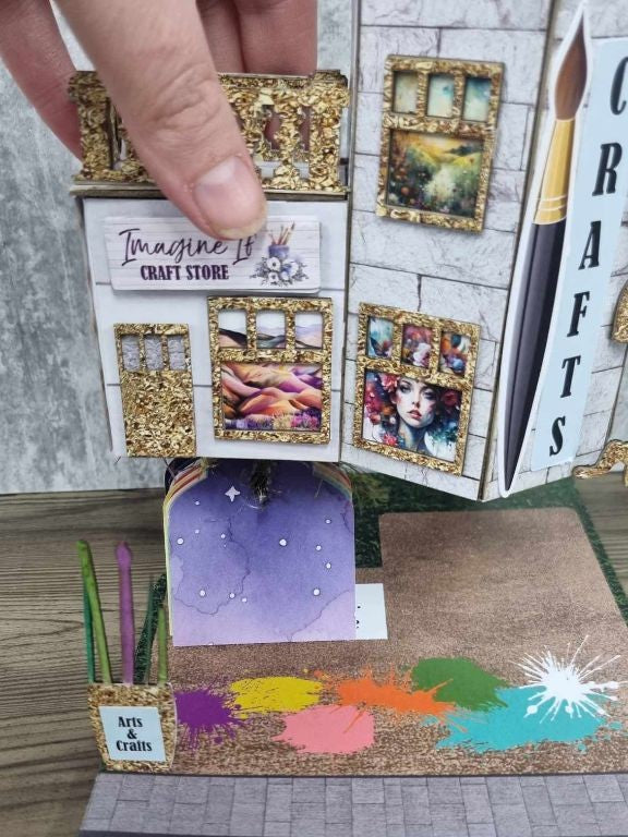 3Quarter Designs Imagine If Craft Store miniature building being lifted to reveal a hidden watercolour galaxy tag book underneath, with a purple night sky tag visible inside the balcony area, Imagine If Craft Store sign, gold glitter ornate window frames, Arts and Crafts sign, coloured pencil die-cuts, and colourful paint splash base board