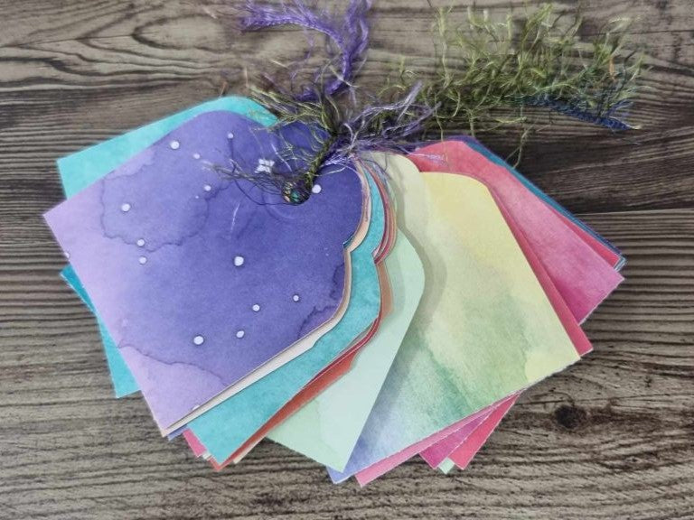 3Quarter Designs Imagine If Craft Store hidden tag book showing a fanned stack of watercolour rainbow tags in purple, teal, coral, yellow, and pink, tied together with purple ribbon and dried botanicals, with a galaxy night sky purple tag on top featuring white dot star details