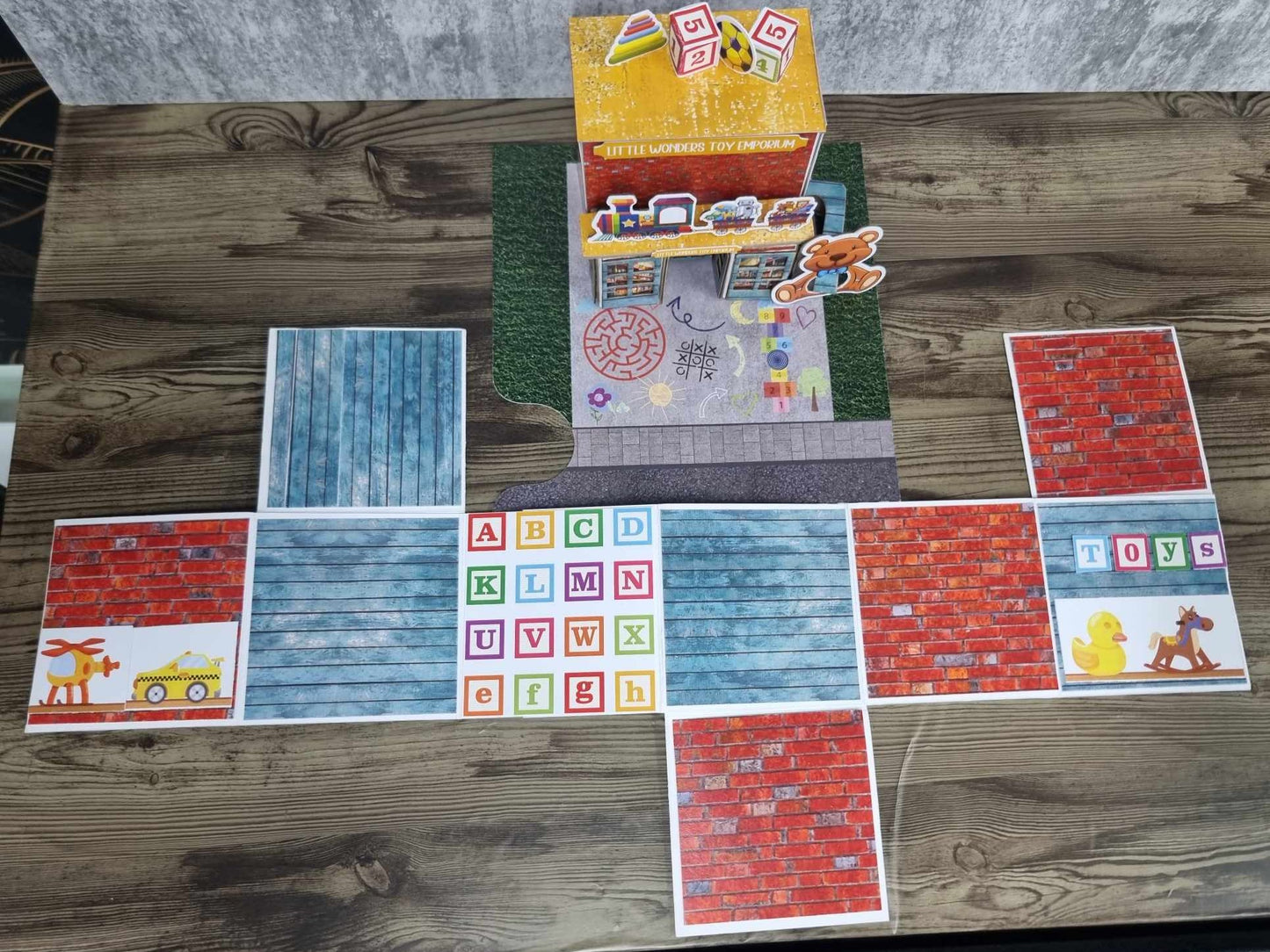 3Quarter Designs Little Wonders Toy Emporium mini album open and laid flat showing red brick and blue timber-look pages, alphabet block grid, and toy die-cuts including rubber duck, rocking horse, and yellow taxi, with assembled toy shop in background
