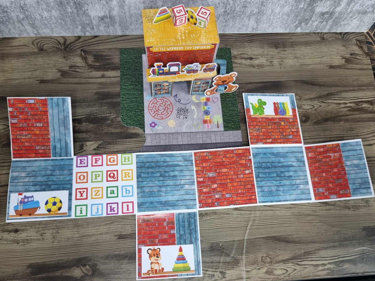 3Quarter Designs Little Wonders Toy Emporium mini album open showing reverse side of pages with red brick and blue timber-look papers, alphabet block grid, and toy die-cuts including toy boat, soccer ball, tiger cub, stacking rings, green dinosaur, and colourful skittles