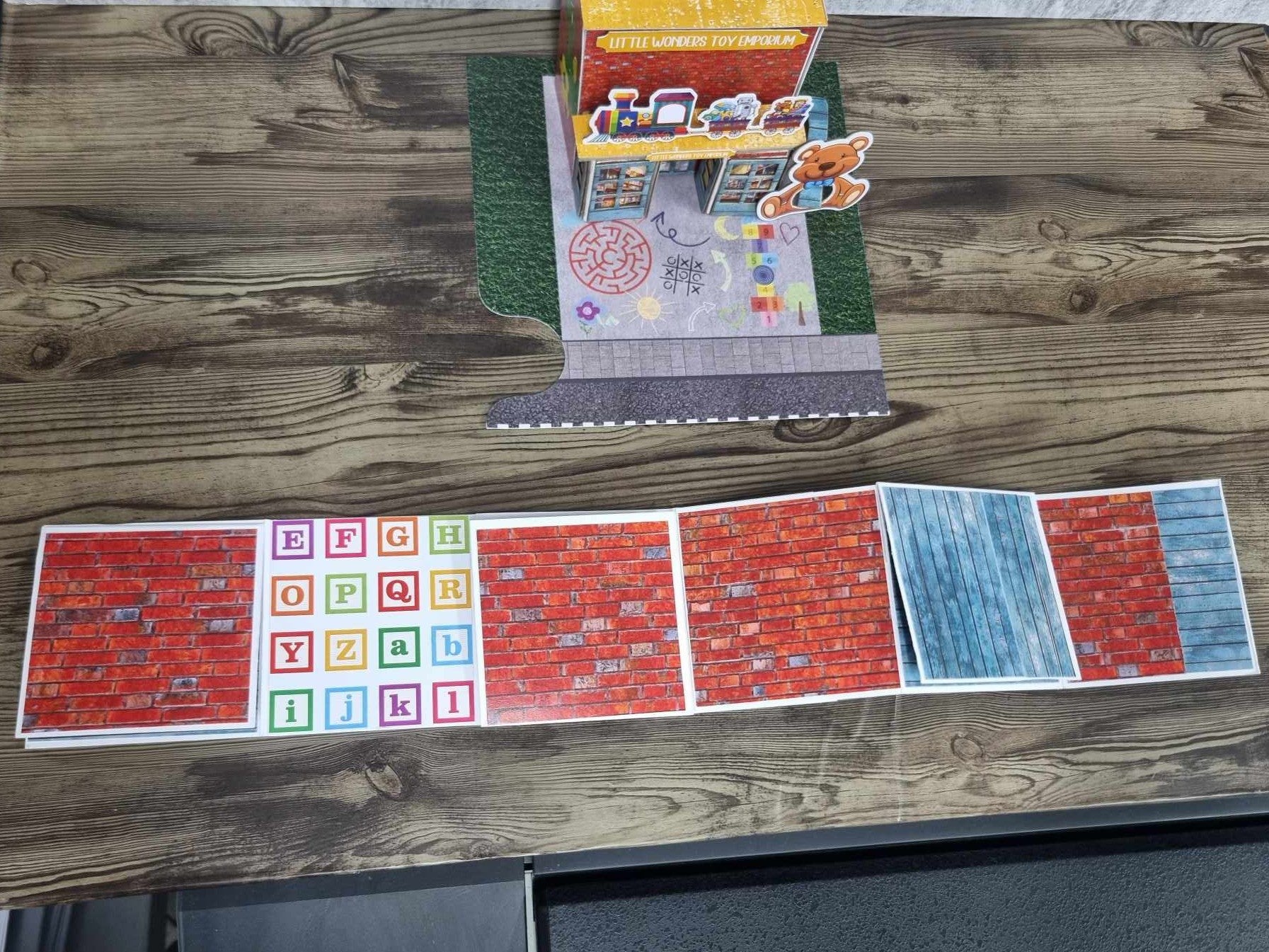 3Quarter Designs Little Wonders Toy Emporium mini album open with top and bottom flaps closed, showing accordion pages with red brick and blue timber-look papers and alphabet block grid, with assembled toy shop in background