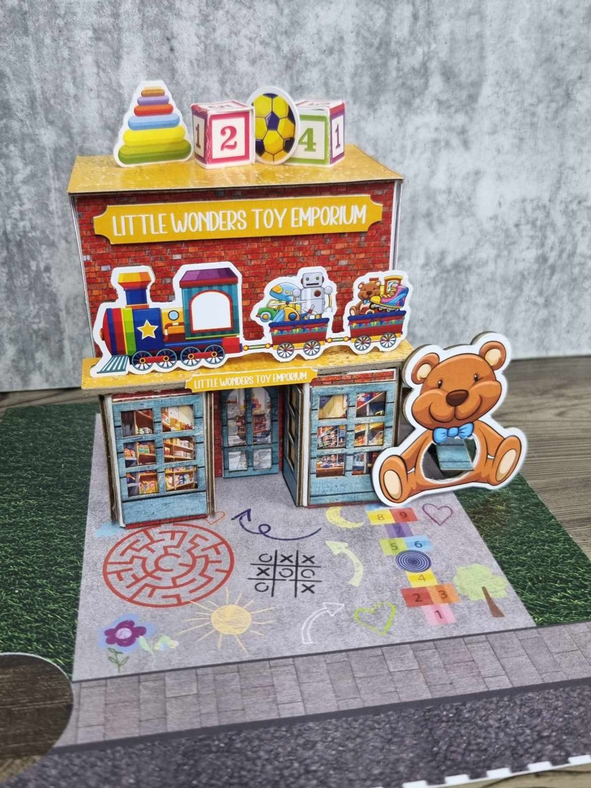 3Quarter Designs Little Wonders Toy Emporium front view showing toy train awning, number block and soccer ball rooftop details, and chalk-art playground base board