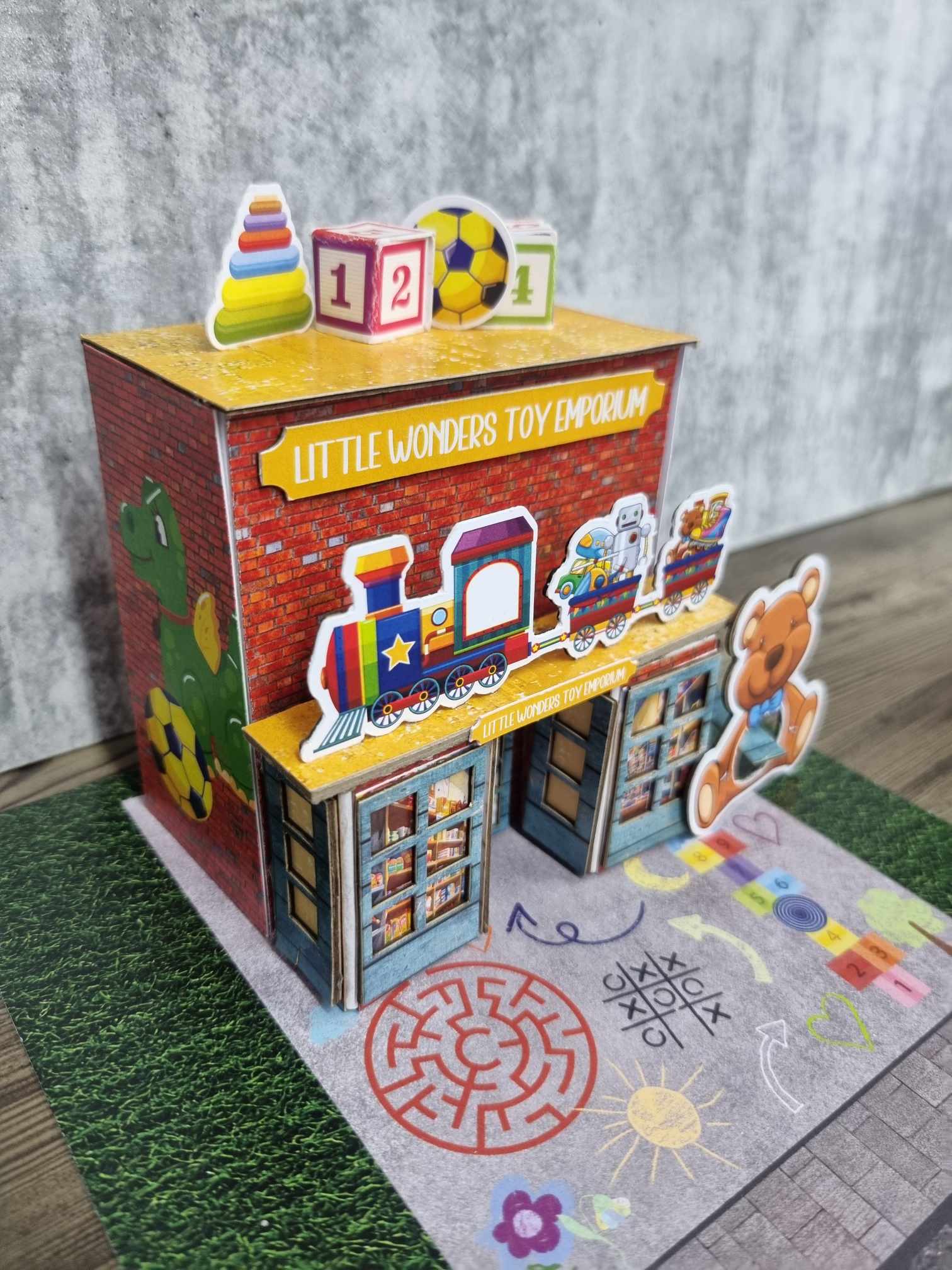 3Quarter Designs Little Wonders Toy Emporium assembled miniature toy shop — angled front view with teddy bear die-cut, colourful toy train awning, and chalk-art playground base board