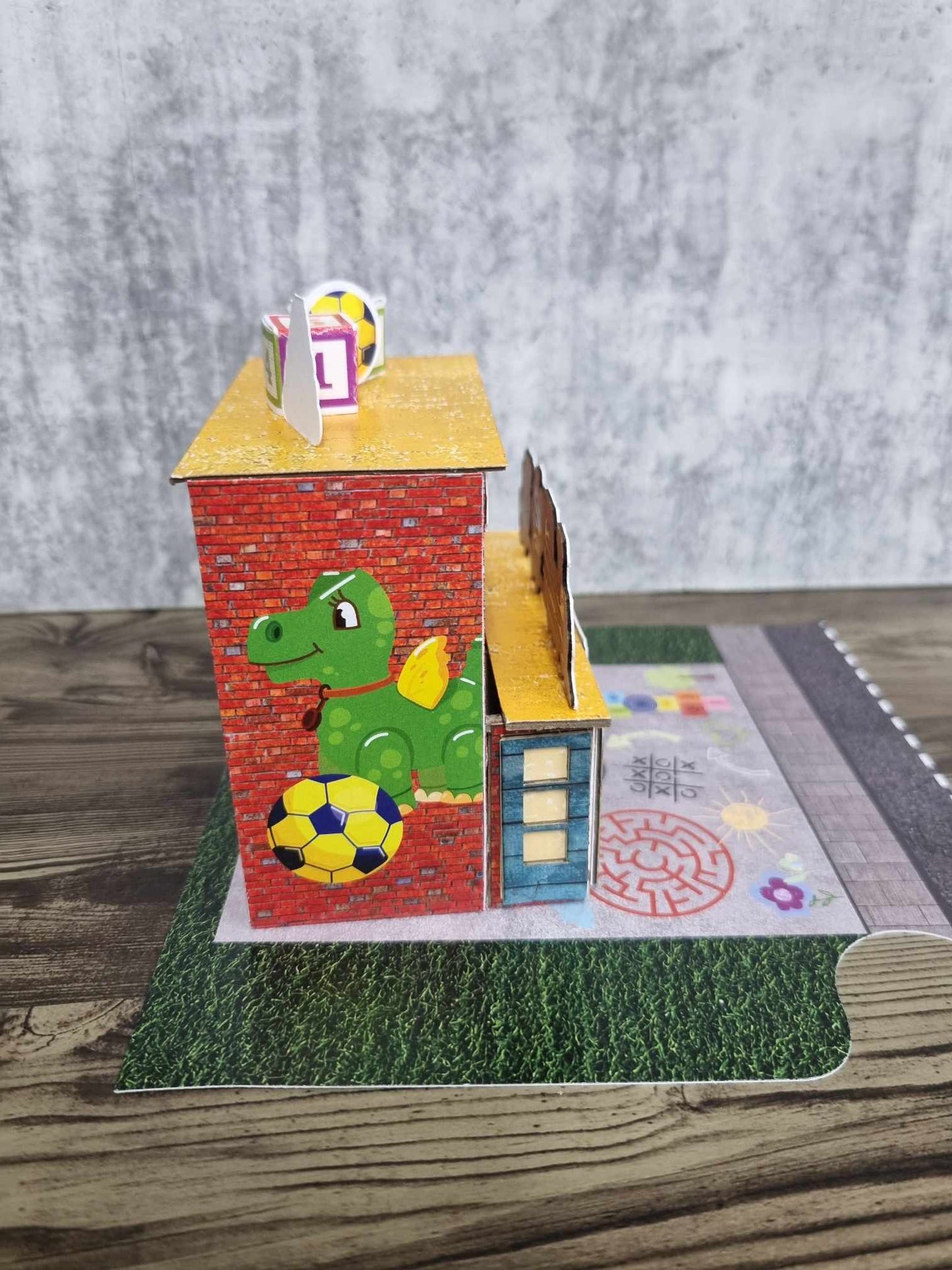 3Quarter Designs Little Wonders Toy Emporium side view showing green dragon die-cut with soccer ball on red brick wall and rooftop number blocks