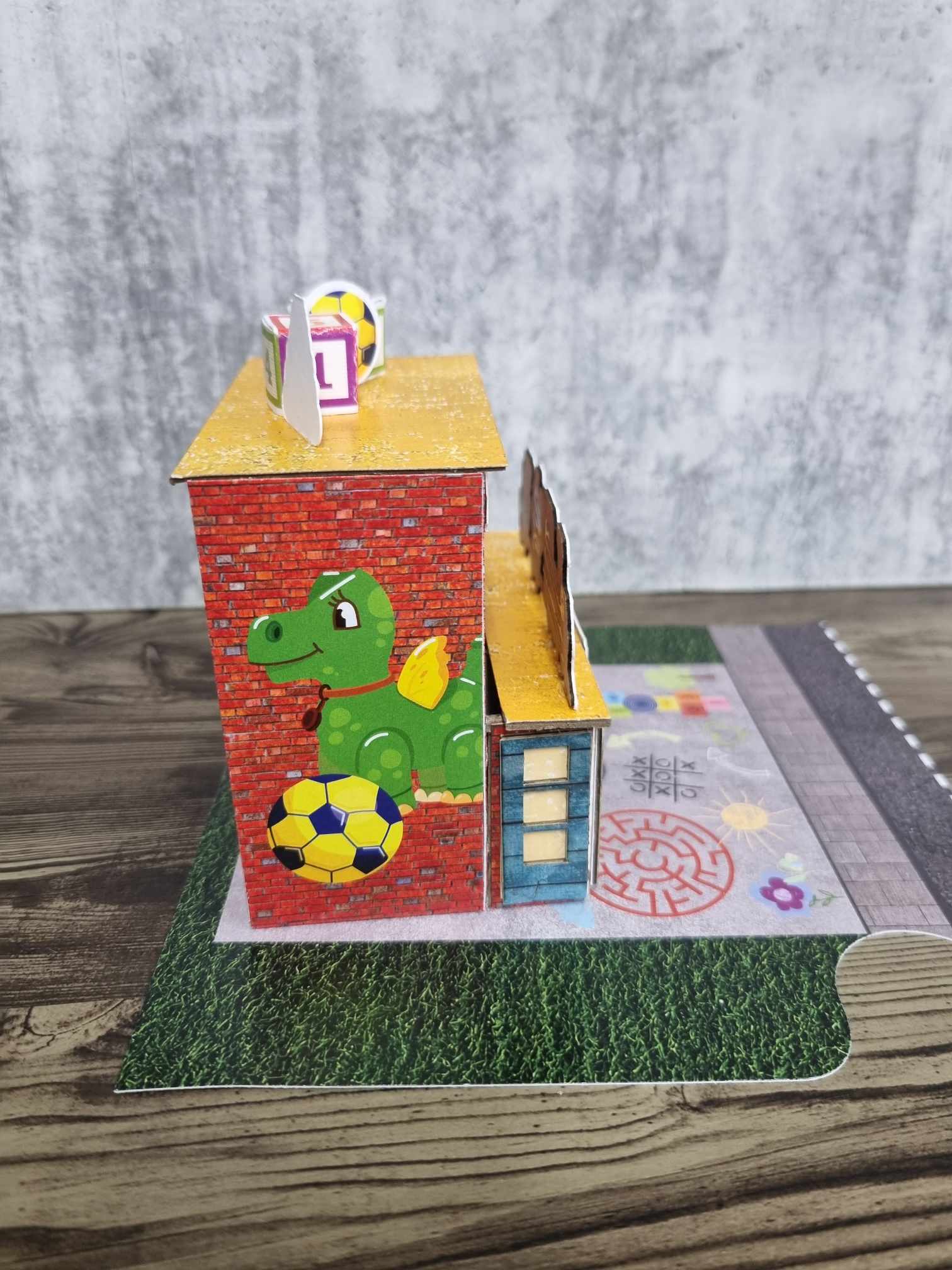 3Quarter Designs Little Wonders Toy Emporium side view showing green dragon die-cut with soccer ball on red brick wall and rooftop number blocks