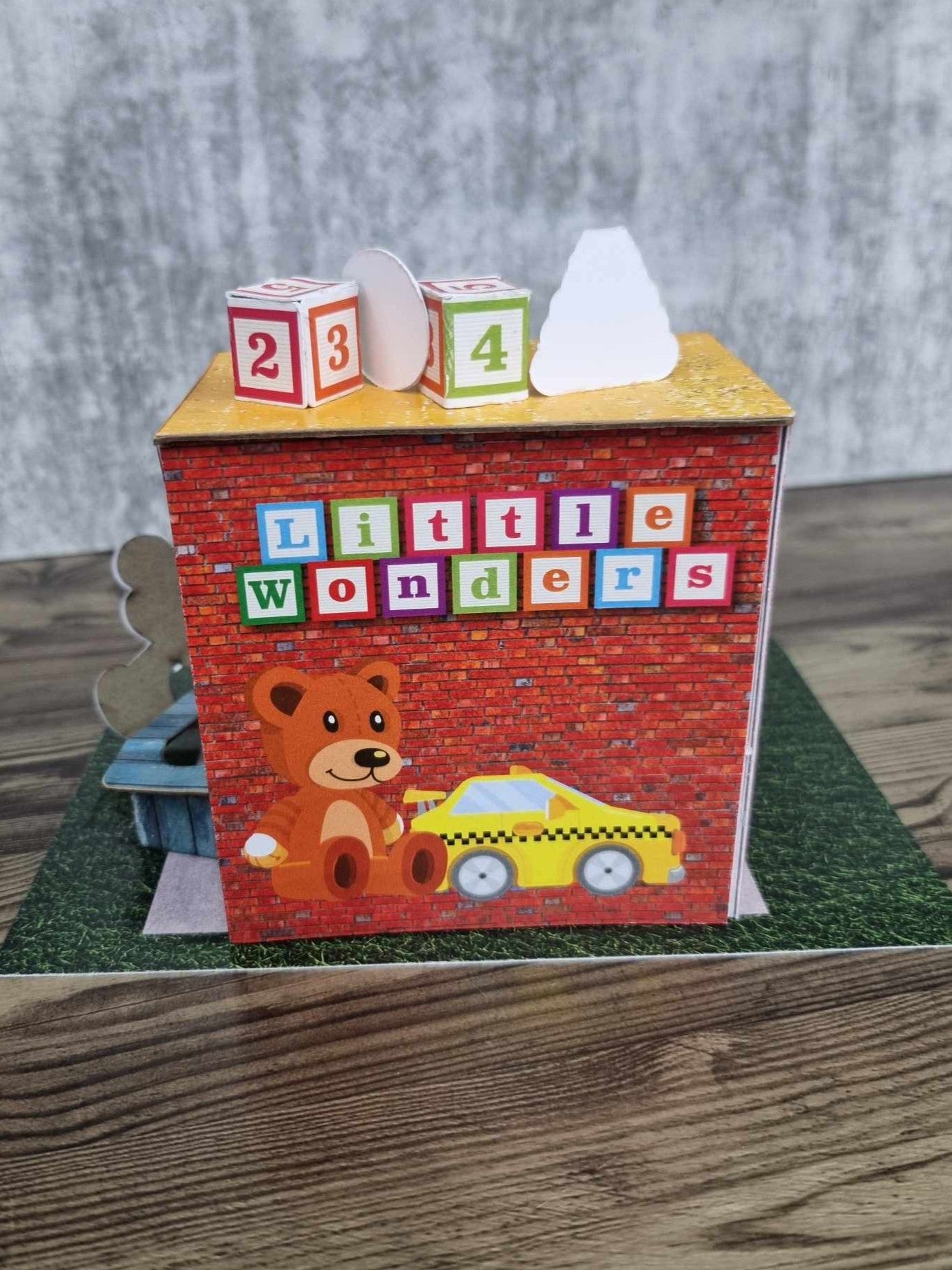 3Quarter Designs Little Wonders Toy Emporium back view showing Little Wonders alphabet block lettering and yellow taxi cab die-cut on red brick wall