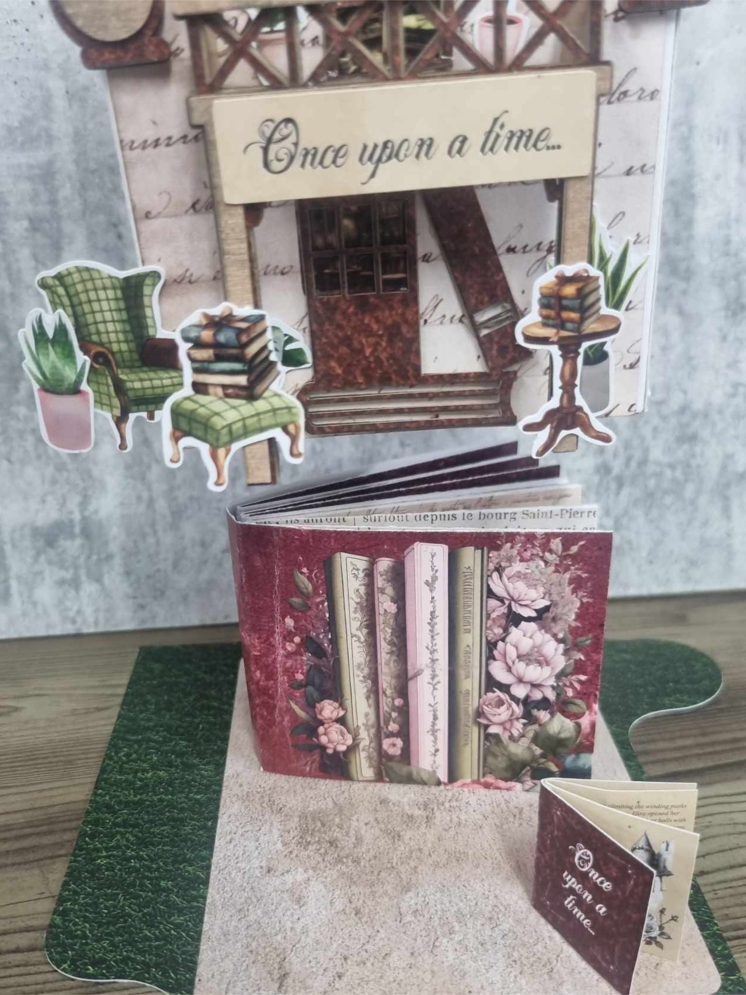 3Quarter Designs Once Upon a Time Book Shop miniature building lifted to reveal the hidden mini album underneath, showing the burgundy floral book spine cover with pink peonies, Once Upon a Time awning, green armchair, stacked books, pedestal stand, and succulent die-cuts on the grass and cobblestone base board