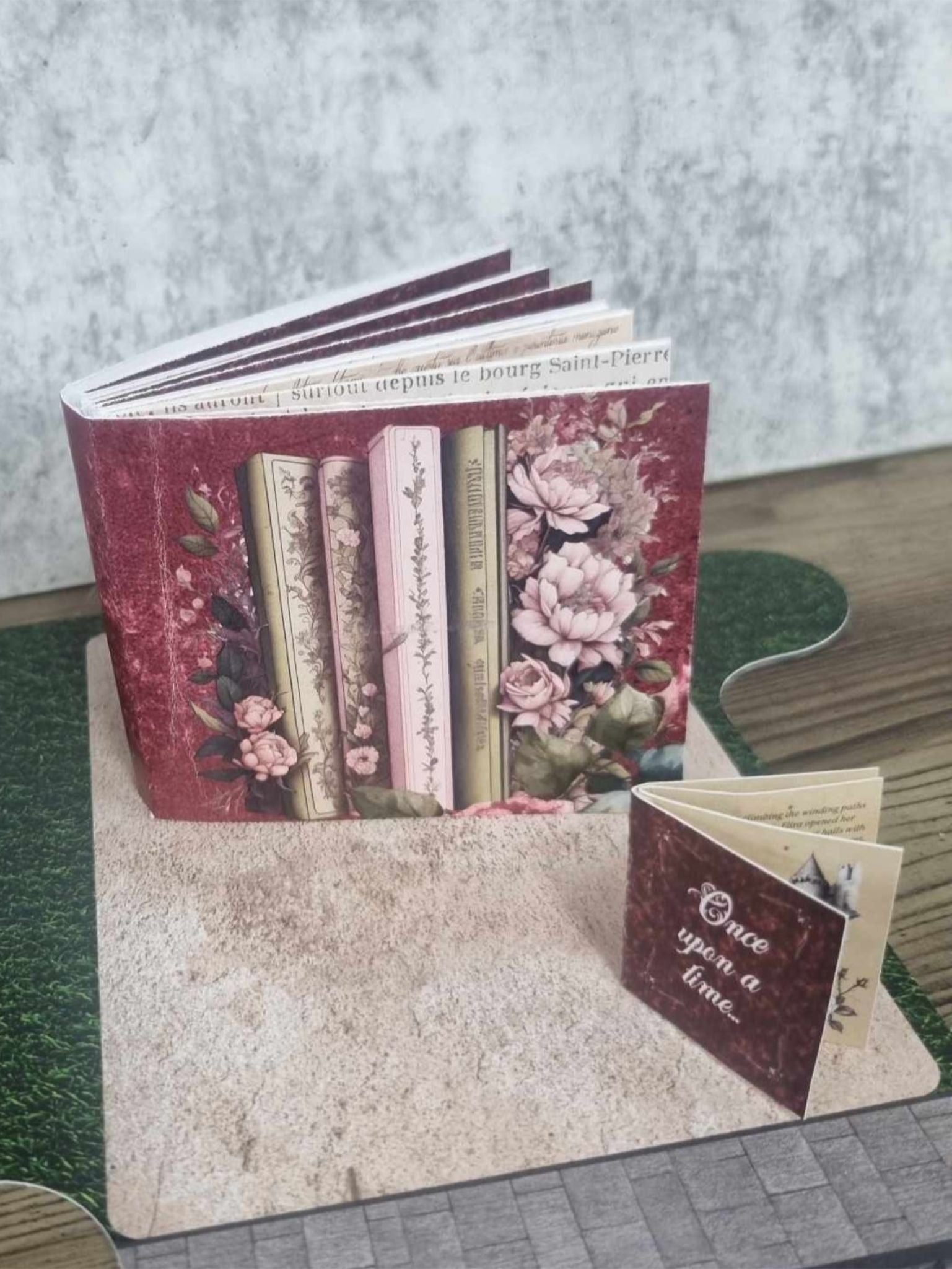 3Quarter Designs Once Upon a Time Book Shop hidden mini album open showing a burgundy floral book spine cover with pink peonies and vintage books, with the small Once Upon a Time companion mini album sitting in front on the cobblestone base board