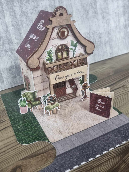 3Quarter Designs Once Upon a Time Book Shop miniature building front view showing aged script facade, burgundy Once Upon a Time roof panel, arched upper window with potted plants, shop awning, reading chair, stacked books, pedestal table, and mini album on cobblestone and grass base board