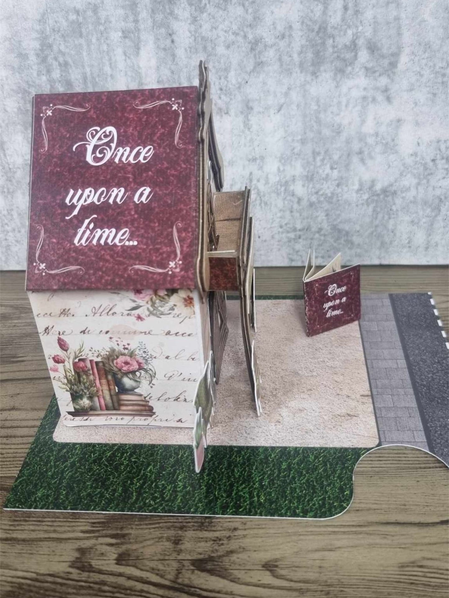 3Quarter Designs Once Upon a Time Book Shop miniature building left side view showing burgundy Once Upon a Time panel with floral frame, aged script lower wall with stacked books and floral vase illustration, exposed chipboard staircase detail, and mini album on base board