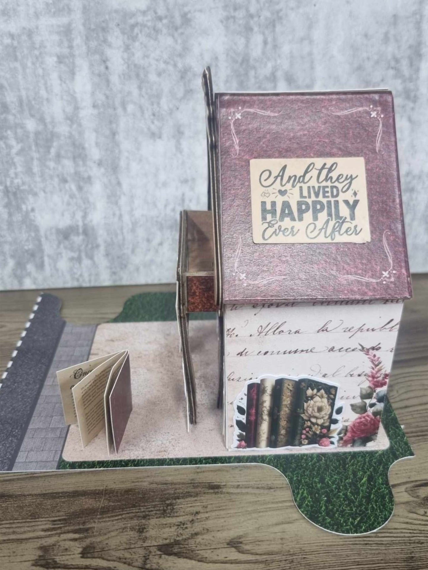 3Quarter Designs Once Upon a Time Book Shop miniature building right side view showing And They Lived Happily Ever After panel, aged script lower wall with floral book stack illustration, exposed chipboard staircase detail, and mini album on cobblestone base board