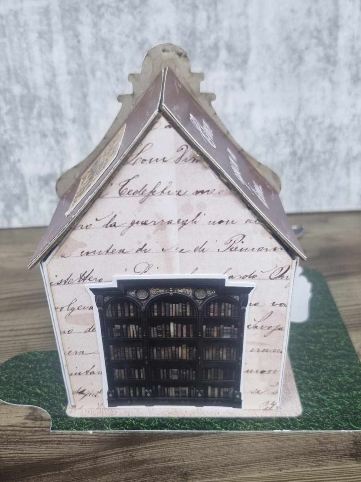 3Quarter Designs Once Upon a Time Book Shop miniature building rear view showing aged script facade with large dark arched bookshelf window filled with rows of miniature books, decorative roofline, and grass jigsaw base board