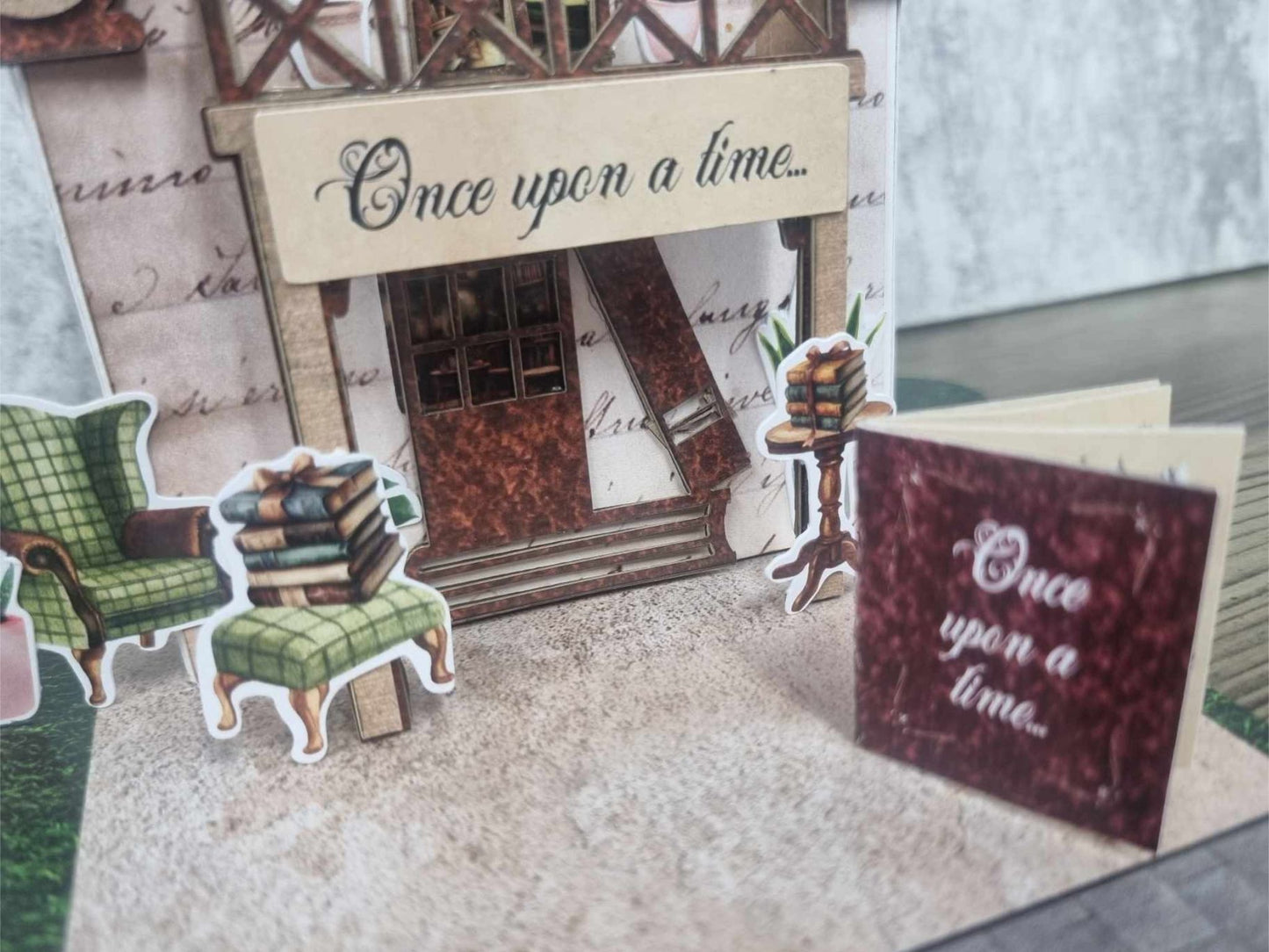  3Quarter Designs Once Upon a Time Book Shop miniature building front view showing Once Upon a Time awning, arched doorway, green armchair and stacked books die-cuts, pedestal book stand, and mini album with burgundy Once Upon a Time cover sitting on the cobblestone base board