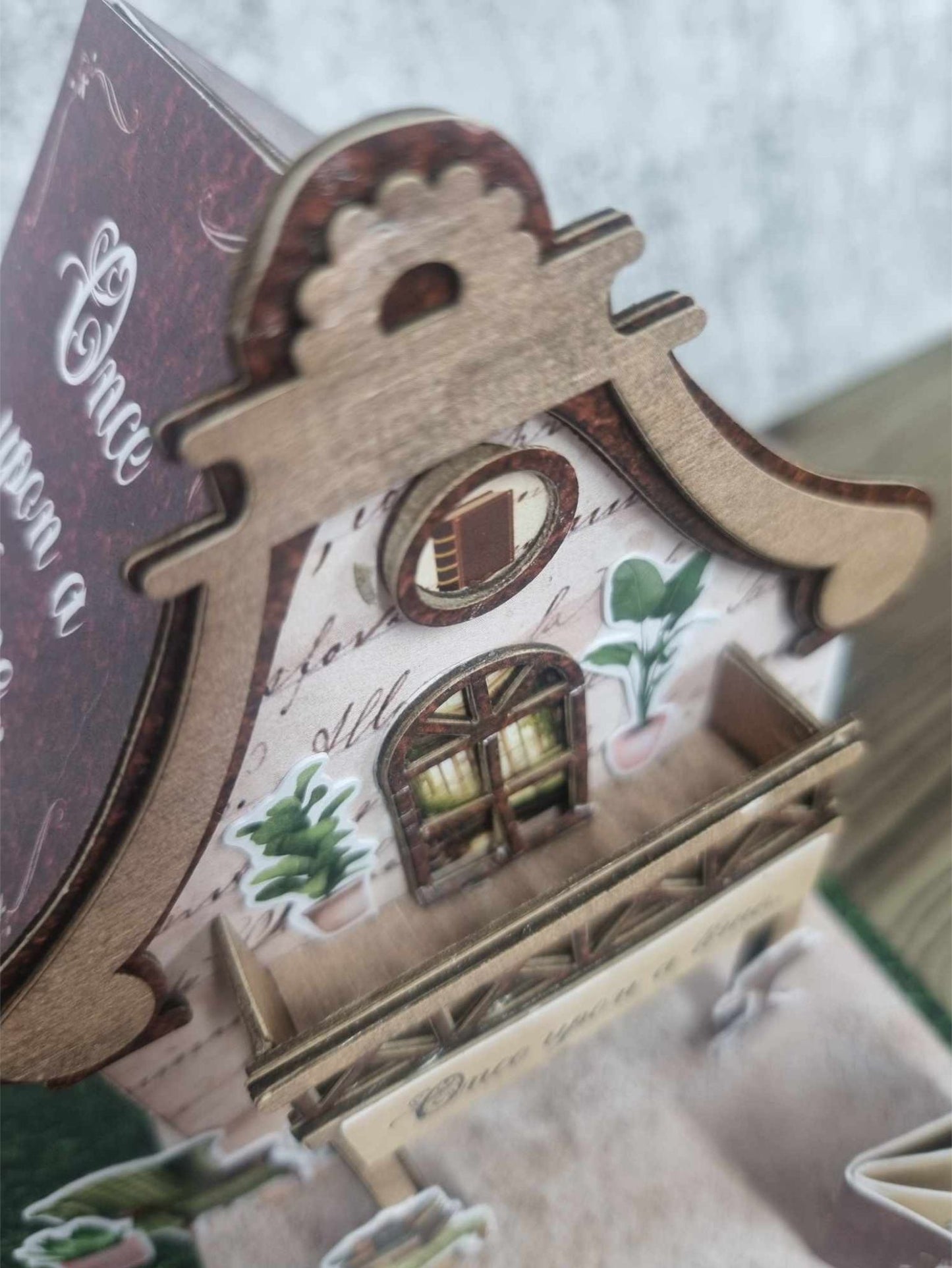 Close-up of 3Quarter Designs Once Upon a Time Book Shop miniature building showing ornate decorative chipboard gable, circular porthole window, arched window with potted plants on aged script and botanical paper, verandah railing, and mini album with Once Upon a Time cover in the foreground