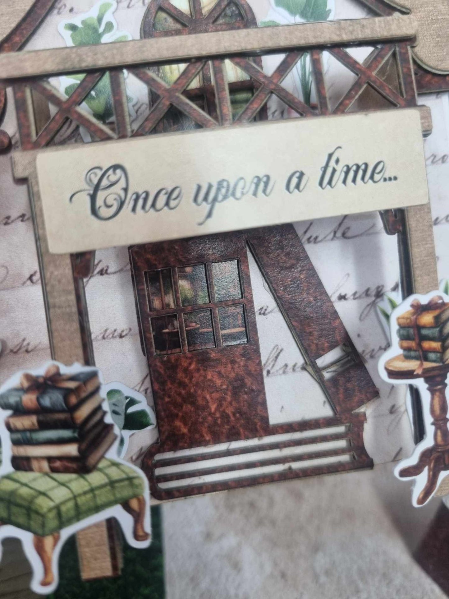 Close-up of 3Quarter Designs Once Upon a Time Book Shop miniature building showing Once Upon a Time awning sign, dark arched doorway with grid window, lattice verandah railing, green armchair die-cut, stacked books die-cut, and pedestal book stand die-cut on cobblestone path