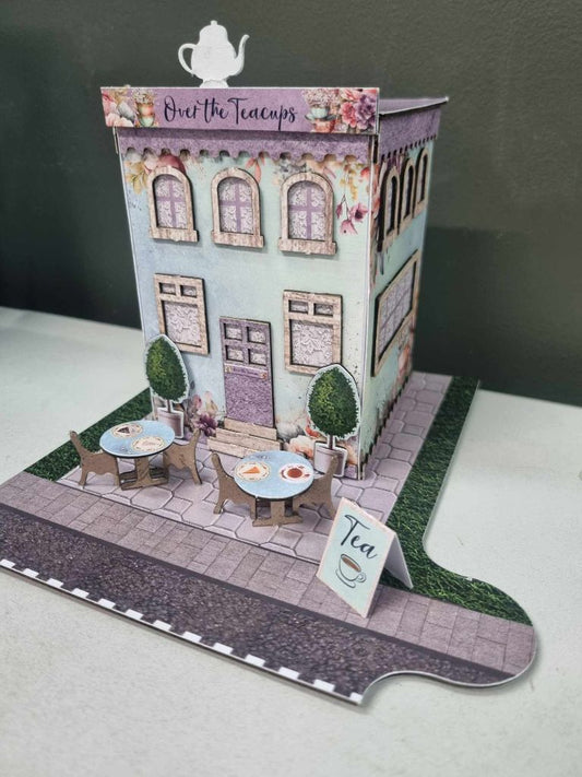 Front view of the Over the Teacups Café with cobblestone terrace, outdoor café tables, chairs, and topiary trees
