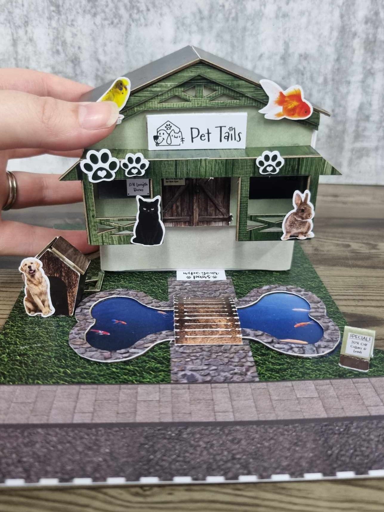 3Quarter Designs Pet Tails Pet Shop being lifted to reveal the paper basket mini album sitting inside the shop, with the bone-shaped pond and die-cut animals visible
