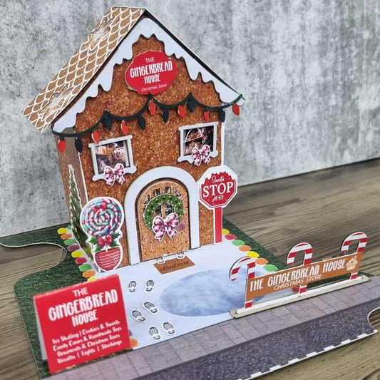 3Quarter Designs Gingerbread House Christmas Store miniature building angled front view showing glittery gingerbread textured walls, scalloped icing roof with die-cut fairy lights, The Gingerbread House Christmas Store sign, arched door with Christmas wreath and pink bow, elf feet die-cut, Merry Christmas doormat, lollipop topiary die-cut, Santa Stop Here sign, candy cane fence, colourful gumdrop border, snow-covered base board with footprints, and Gingerbread House menu sign on grass jigsaw base board