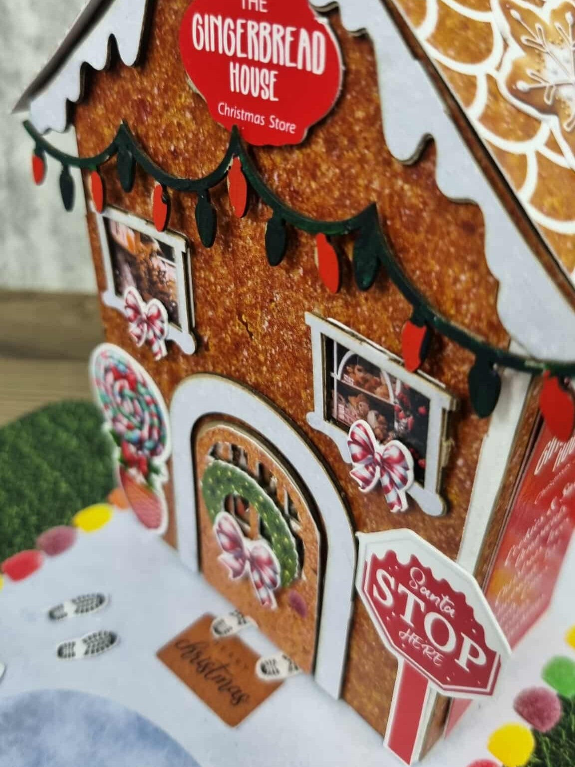 Close-up of 3Quarter Designs Gingerbread House Christmas Store miniature building showing glittery gingerbread texture, die-cut fairy light garland, The Gingerbread House Christmas Store sign, two photo windows with Christmas scene prints and pink candy bow die-cuts, lollipop topiary die-cut, arched door with Christmas wreath and pink bow, elf feet die-cut, Santa Stop Here sign, and colourful gumdrop border on snow base board