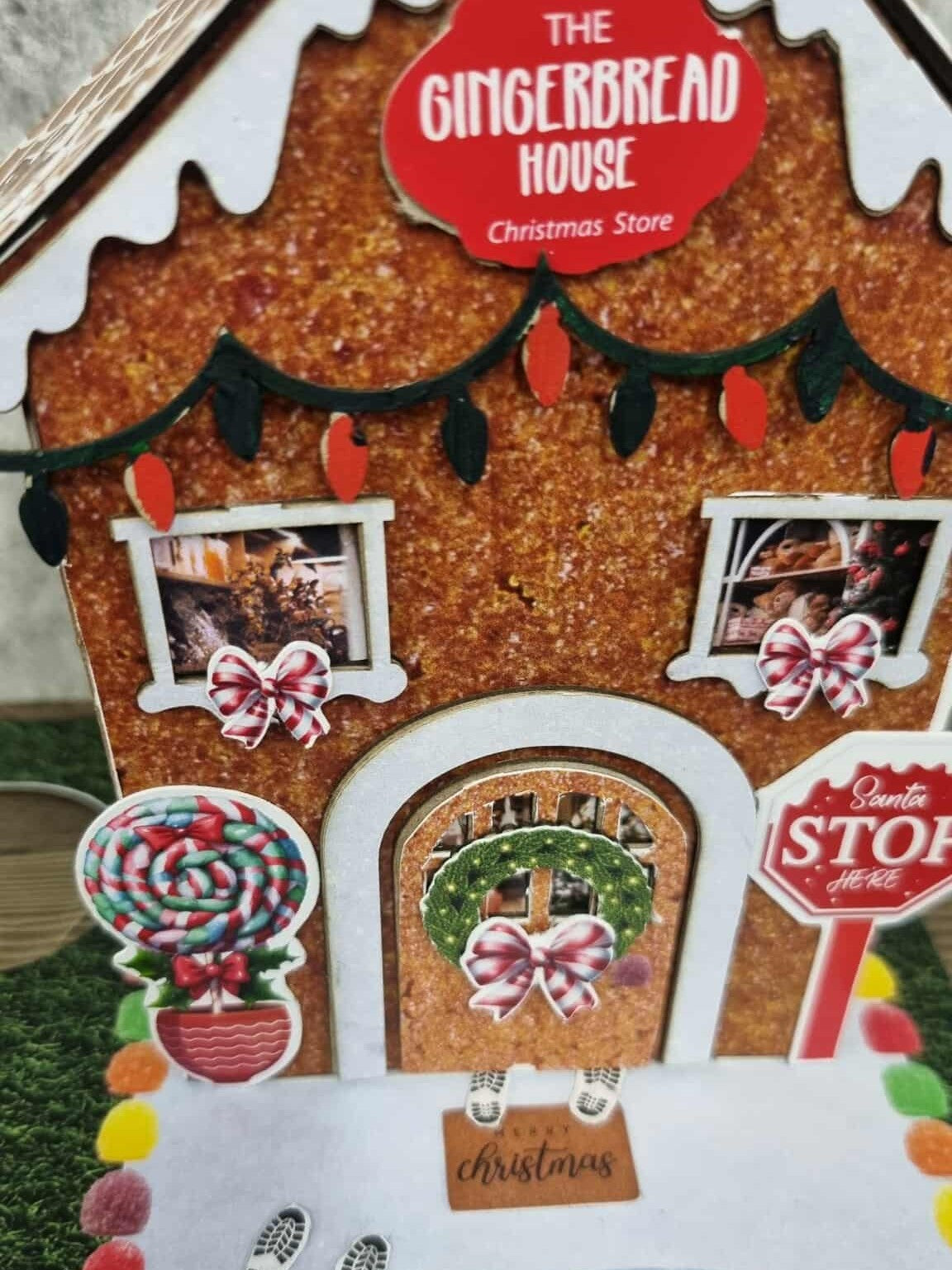 Close-up of 3Quarter Designs Gingerbread House Christmas Store miniature building front showing glittery gingerbread texture, die-cut fairy light garland, two photo windows with Christmas scene prints and pink candy bow die-cuts, lollipop topiary die-cut, arched door with Christmas wreath and pink bow, elf feet die-cut, Merry Christmas doormat, Santa Stop Here sign, and colourful gumdrop border on snow base board