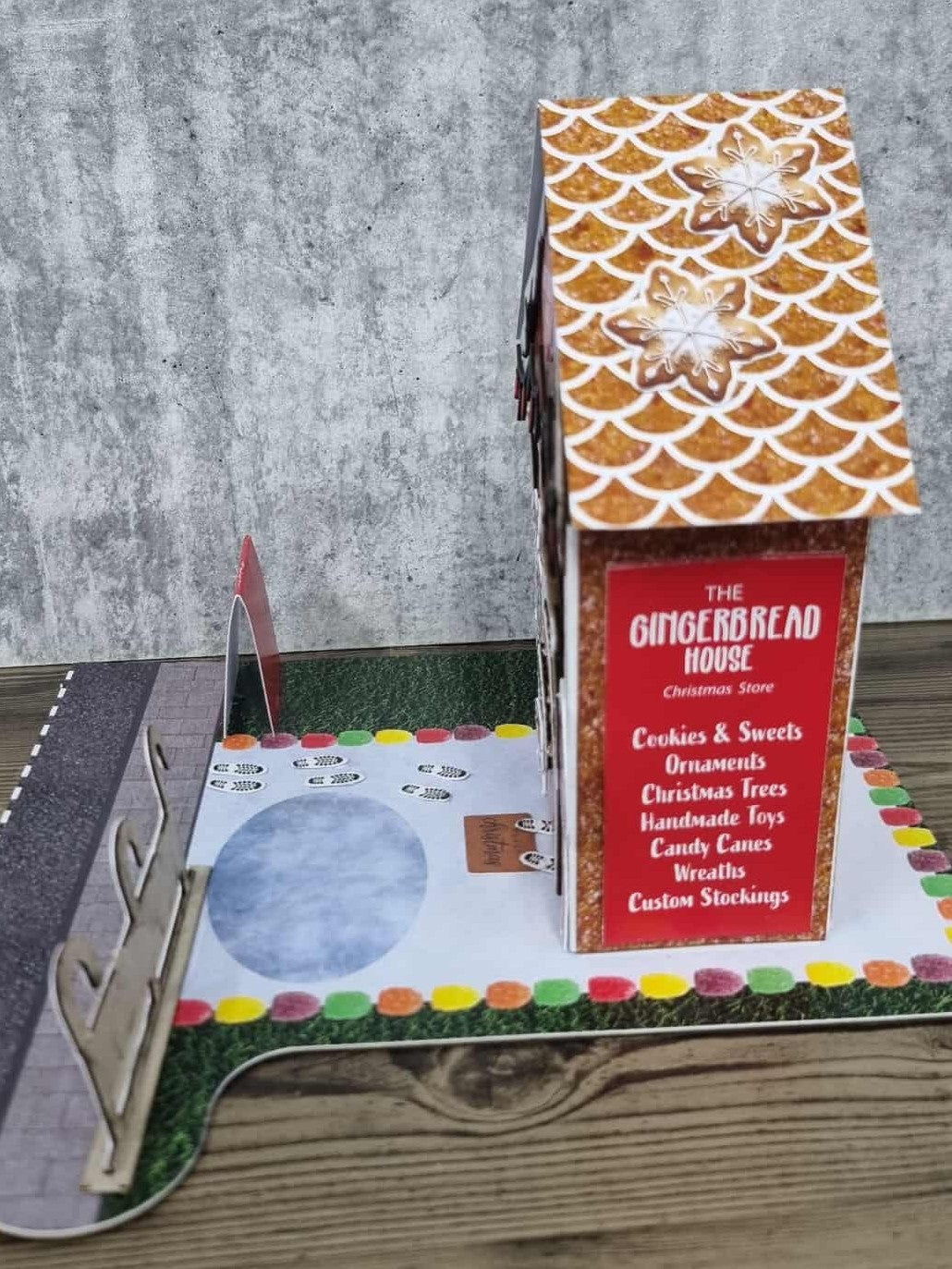 3Quarter Designs Gingerbread House Christmas Store miniature building right side view showing glittery gingerbread textured wall, snowflake cookie scalloped roof tiles, The Gingerbread House Christmas Store red menu sign listing Cookies and Sweets, Ornaments, Christmas Trees, Handmade Toys, Candy Canes, Wreaths, and Custom Stockings, chipboard reindeer fence, snow base board with footprints, and colourful gumdrop border on grass jigsaw base board