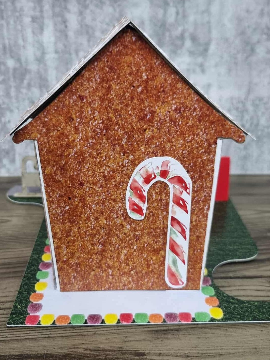 3Quarter Designs Gingerbread House Christmas Store miniature building rear view showing glittery gingerbread textured back wall, snowflake cookie scalloped roof tiles, large candy cane die-cut centred on the back wall, and colourful gumdrop border on grass jigsaw base board