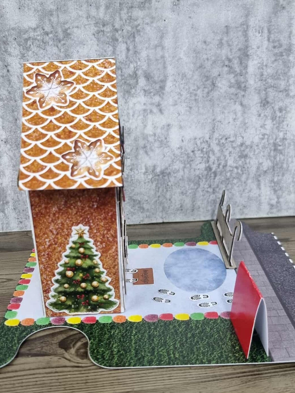 3Quarter Designs Gingerbread House Christmas Store miniature building left side view showing glittery gingerbread textured wall, snowflake cookie scalloped roof tiles, decorated Christmas tree die-cut, chipboard reindeer fence on snow base board, and colourful gumdrop border on grass jigsaw base board