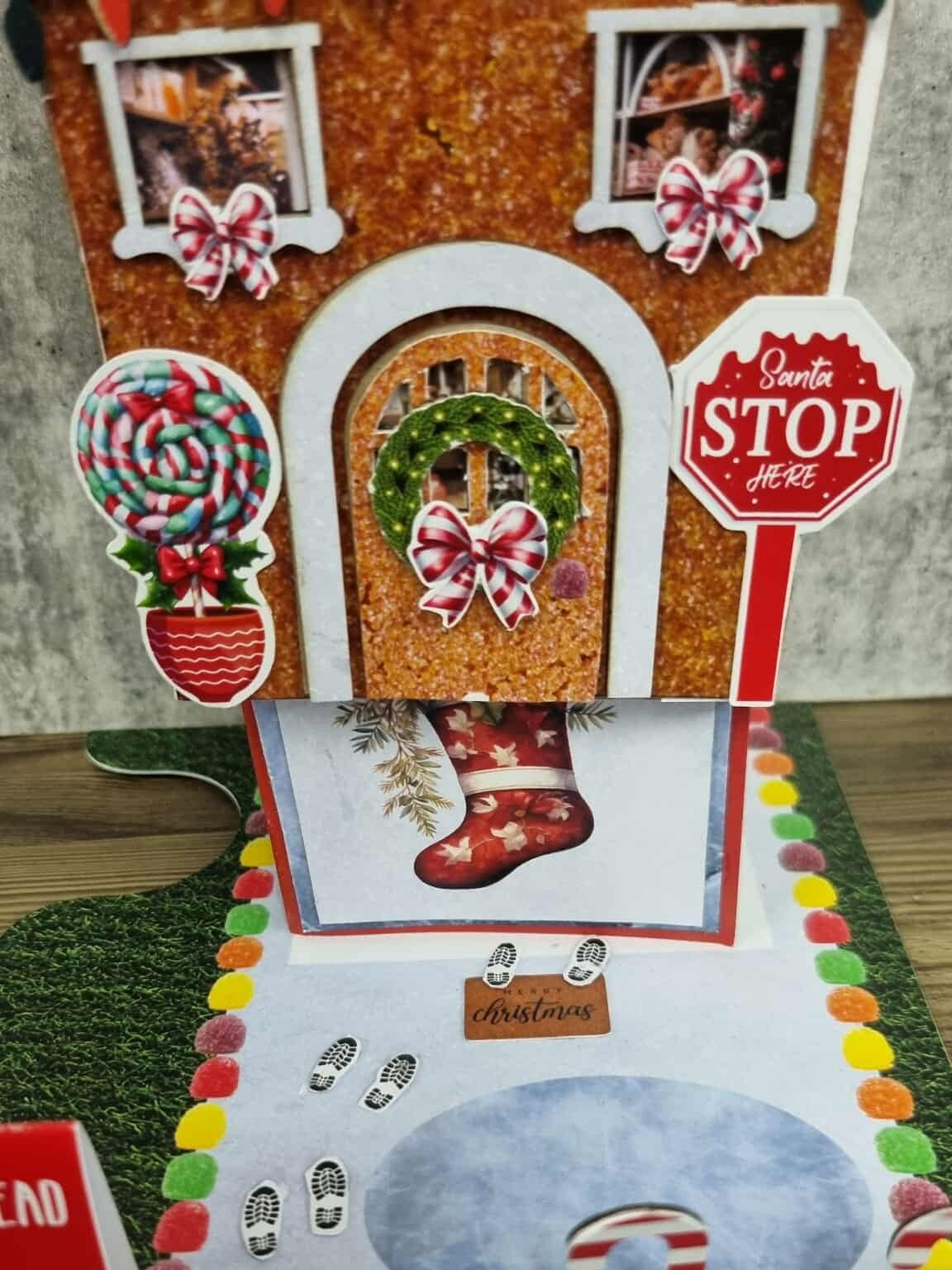 3Quarter Designs Gingerbread House Christmas Store miniature building being lifted to reveal a hidden Christmas stocking card underneath, showing lollipop topiary die-cut, photo windows with candy bow die-cuts, Christmas wreath with pink bow, Santa Stop Here sign, Merry Christmas doormat, snow base board with footprints, and colourful gumdrop border
