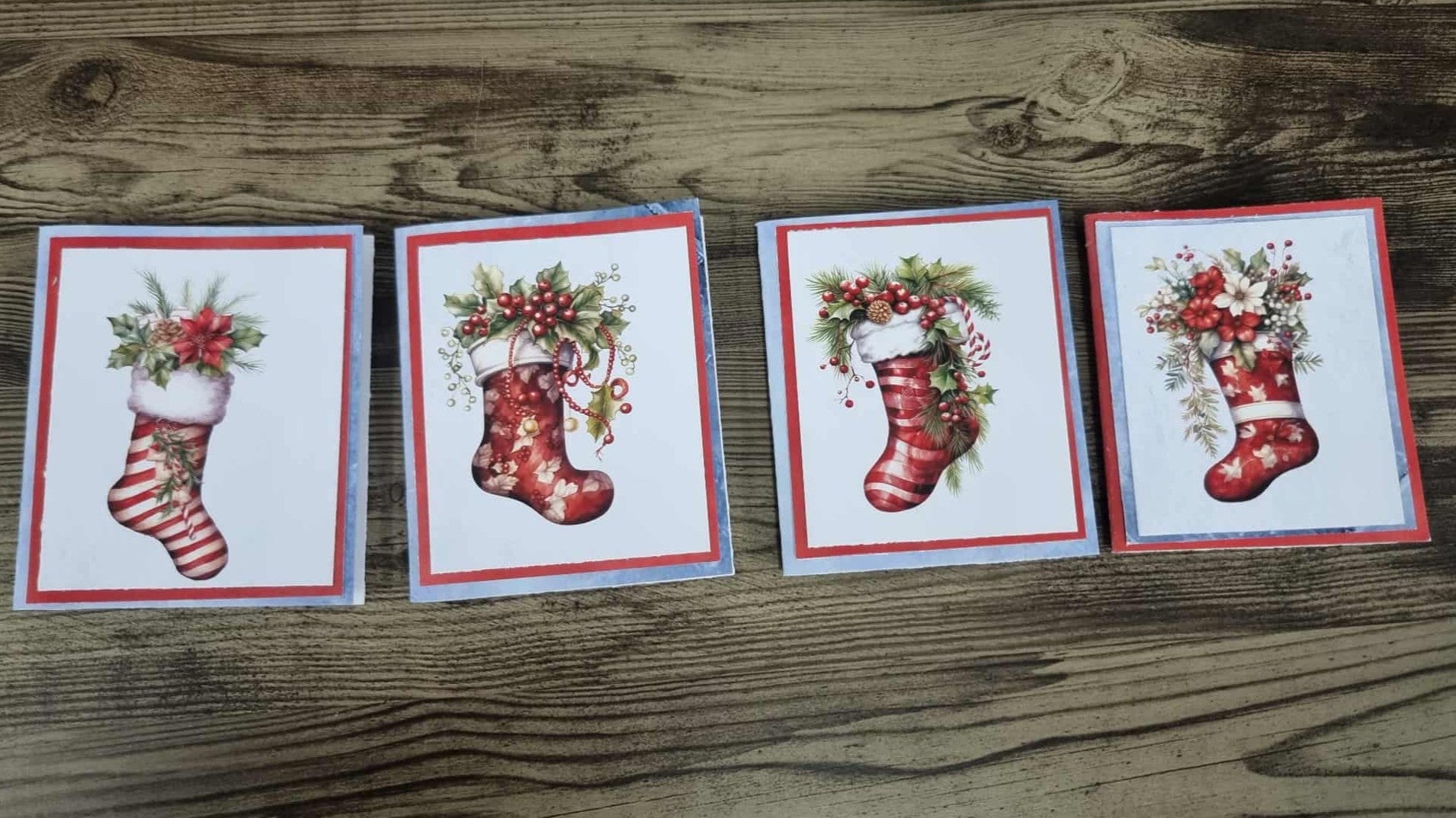 Set of four 3Quarter Designs Gingerbread House Christmas Store hidden Christmas cards laid out on a timber surface, each featuring a different watercolour Christmas stocking design decorated with holly, red berries, pine sprigs, poinsettia blooms, and candy canes on a white card with red and blue layered mat